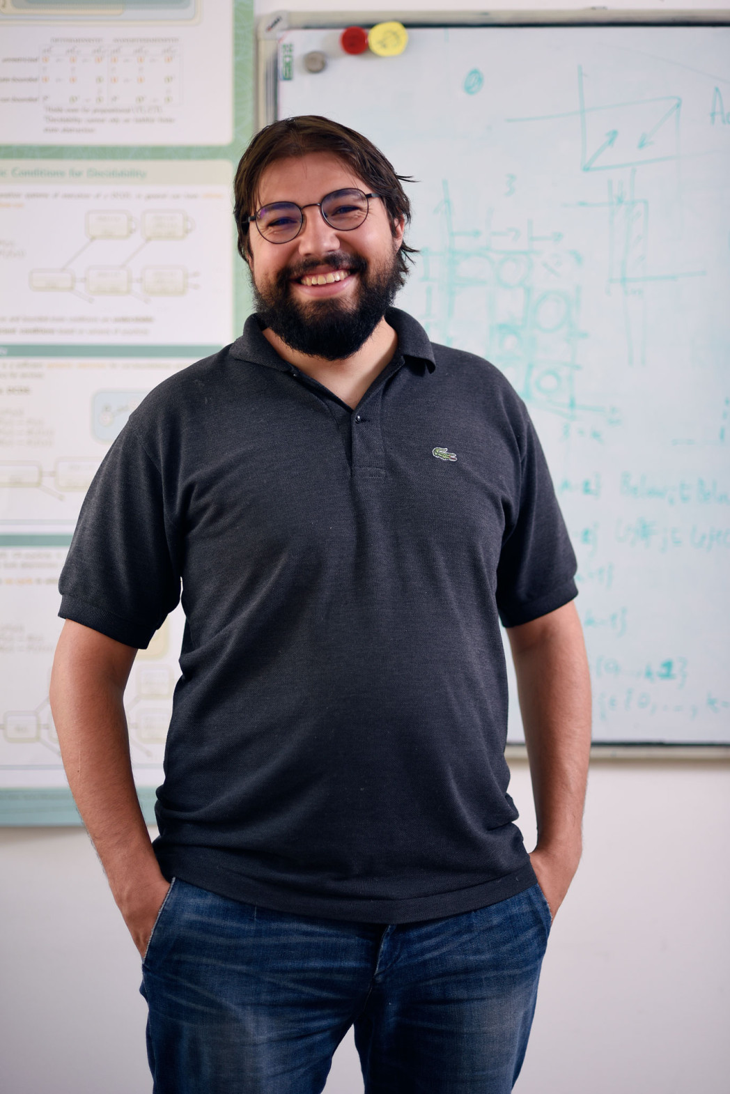 Uomo con barba e occhiali sorride davanti a una lavagna piena di schemi tecnici.