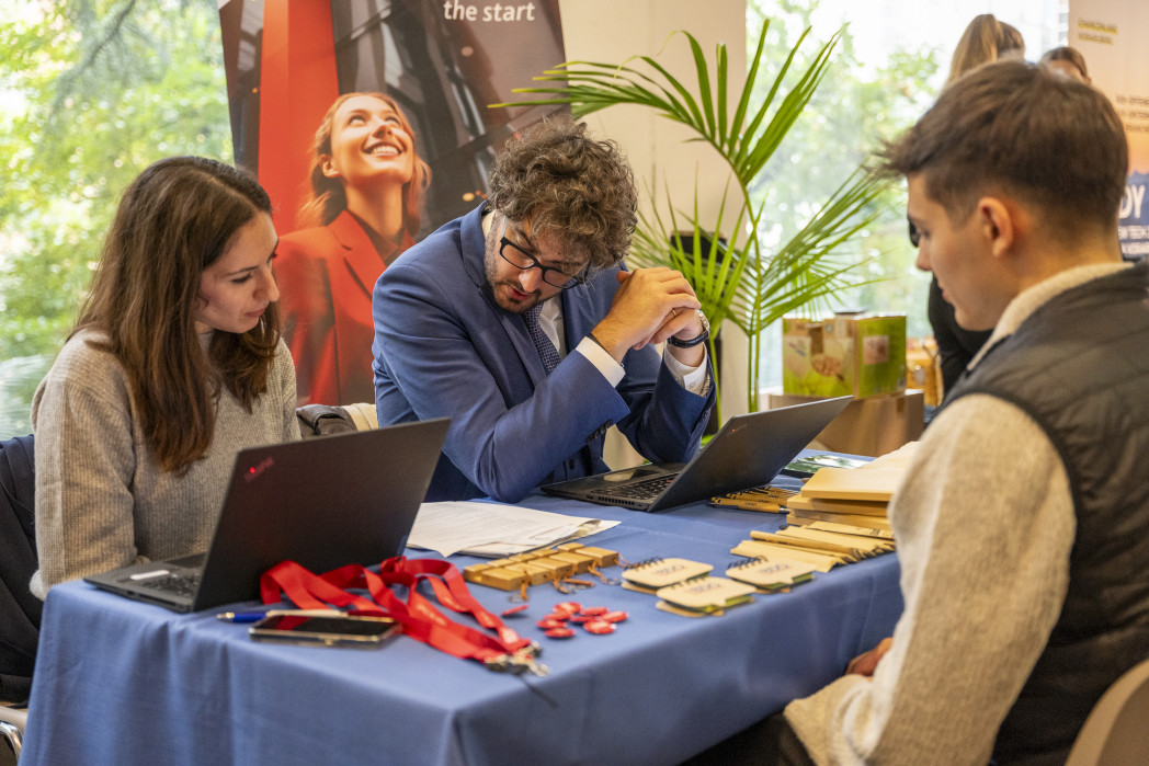 Drei Personen sitzen an einem Tisch: eine Frau und ein Mann schauen in ihrem Laptop. Ihnen sitzt ein anderer Mann gegenüber und schaut sie an.