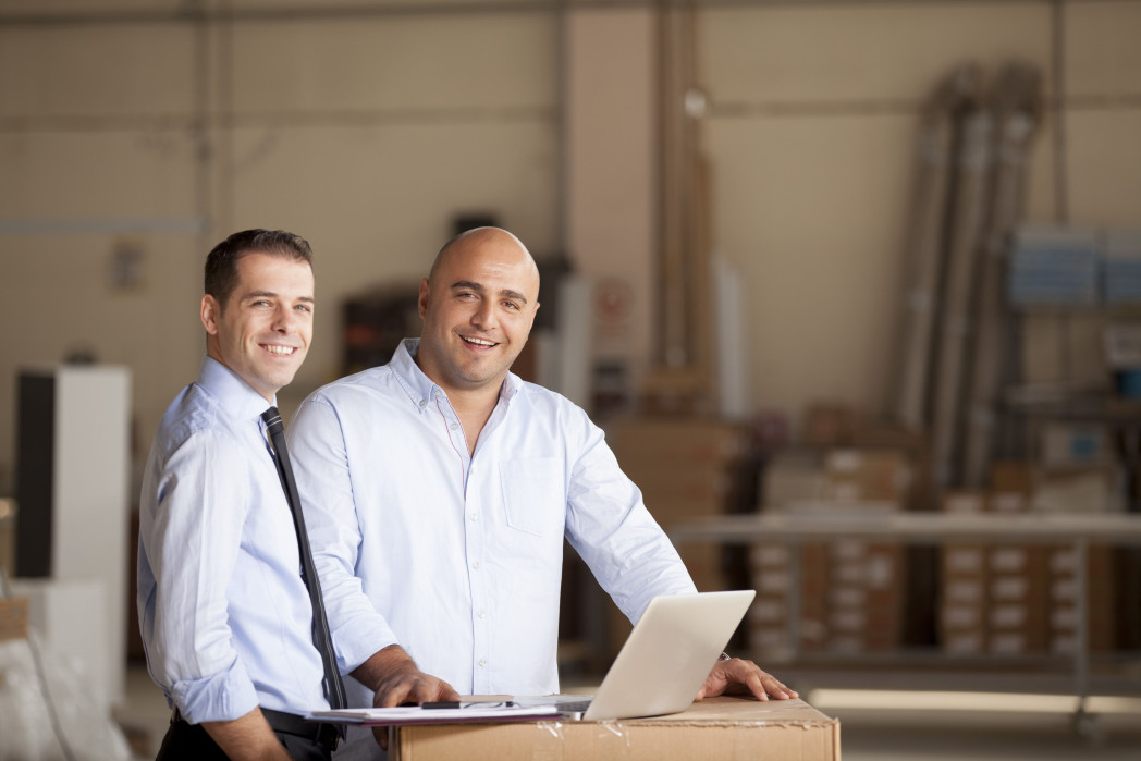 Due uomini sorridenti in un magazzino, uno in camicia e cravatta, l'altro in camicia chiara, con un laptop su una scatola.