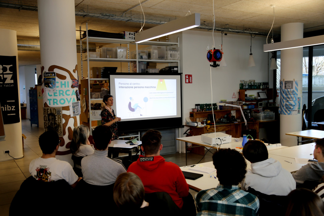 Acht Schüler und Schülerinnen sitzen in einem Klassenzimmer vor einem Bildschirm. Eine Person steht neben dem Bildschirm.