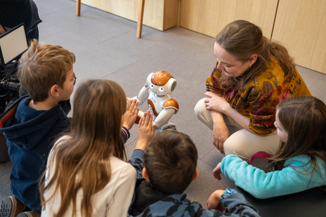 Donna seduta a terra con bambini, al centro un piccolo robot umanoide.