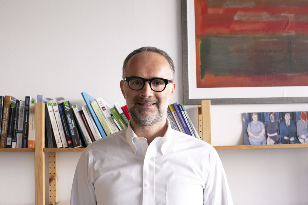 Uomo con occhiali e camicia bianca sorride davanti a una libreria con libri e una cornice con foto e dipinto.