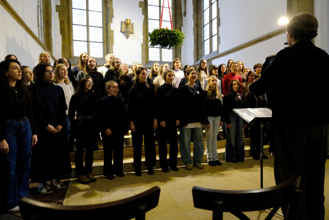 A choir stands at the front of a church, singing together under tall arched windows.