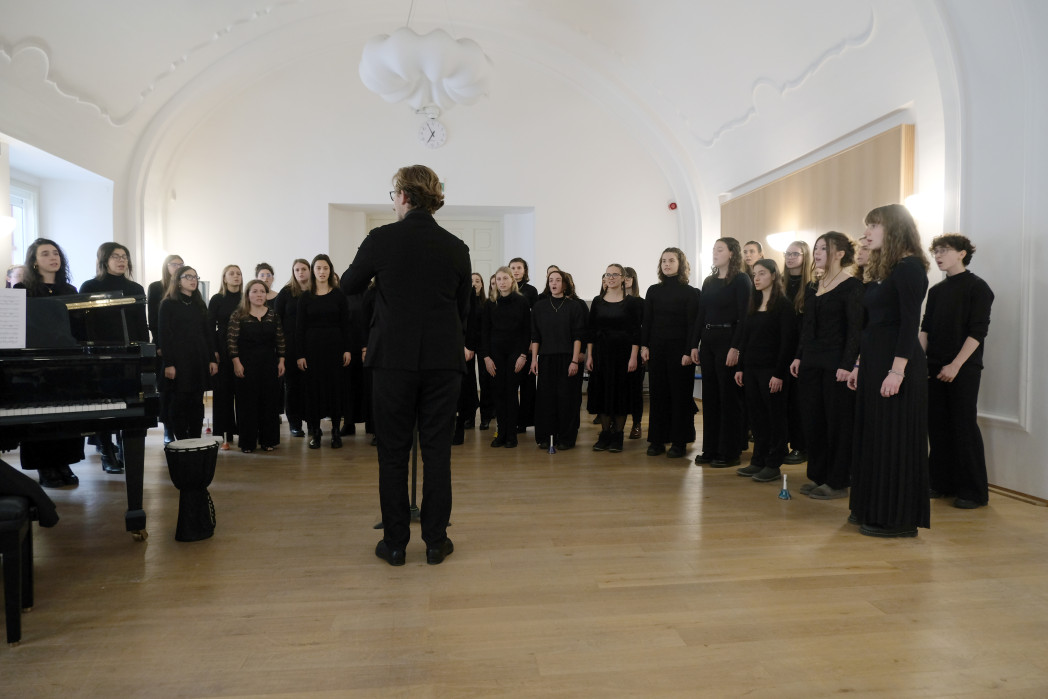 A university choir dressed in black performs in a bright concert hall, led by a conductor standing in front of the singers.