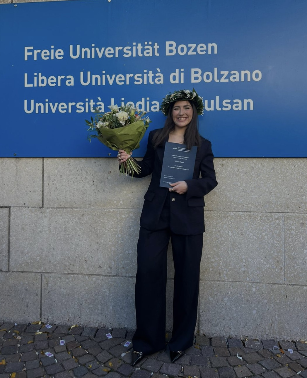Eine Absolventin steht vor dem Schild der Freien Universität Bozen, hält Blumen und ihre Abschlussarbeit und trägt einen traditionellen Lorbeerkranz.