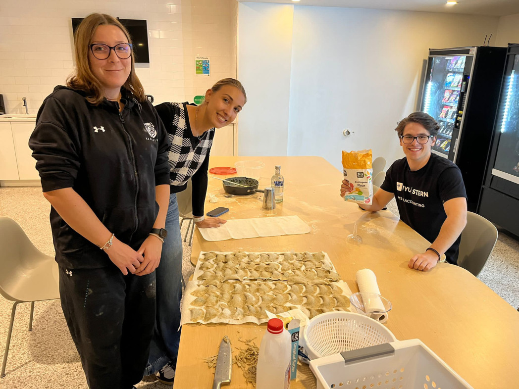 Three people smiling while preparing dumplings together at a table in a kitchen or common area.