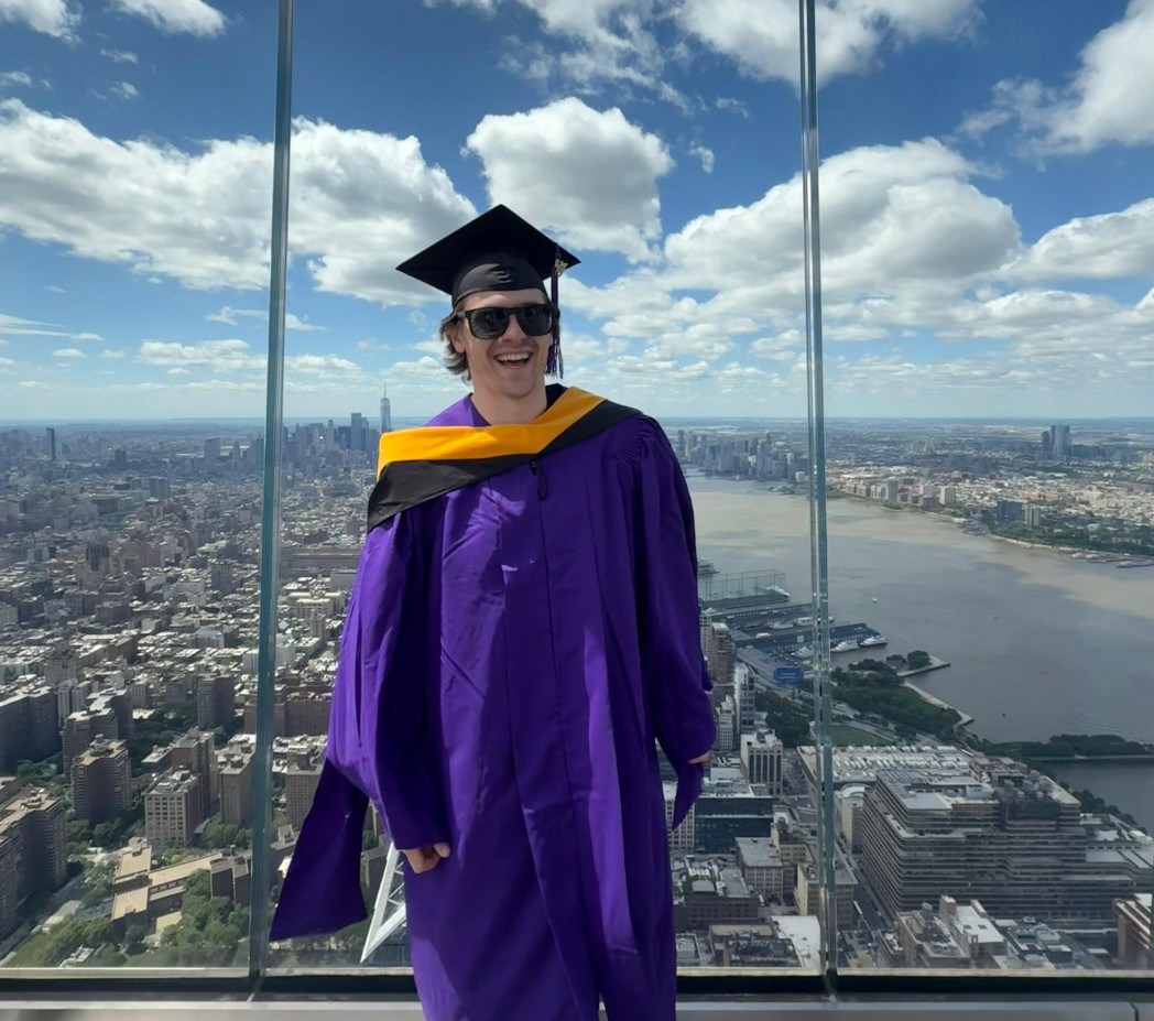 Person wearing a purple graduation gown and cap, smiling in front of a city skyline from a high observation deck.