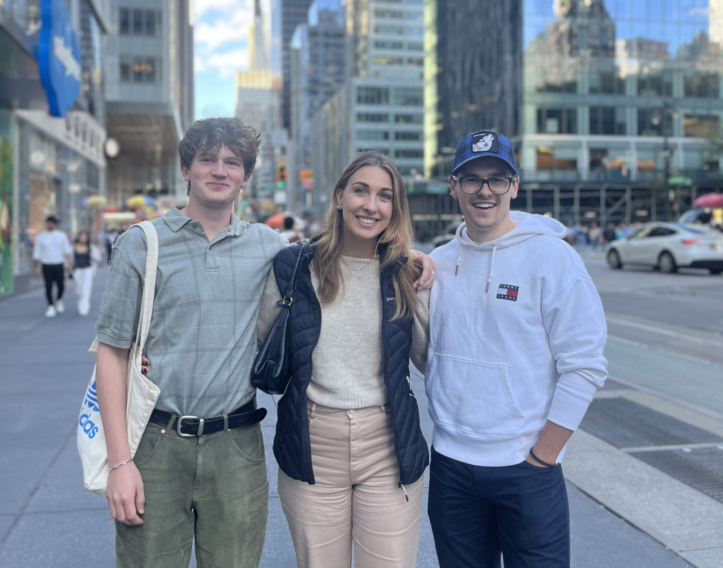 Three friends smiling and posing together on a busy city street surrounded by tall buildings.