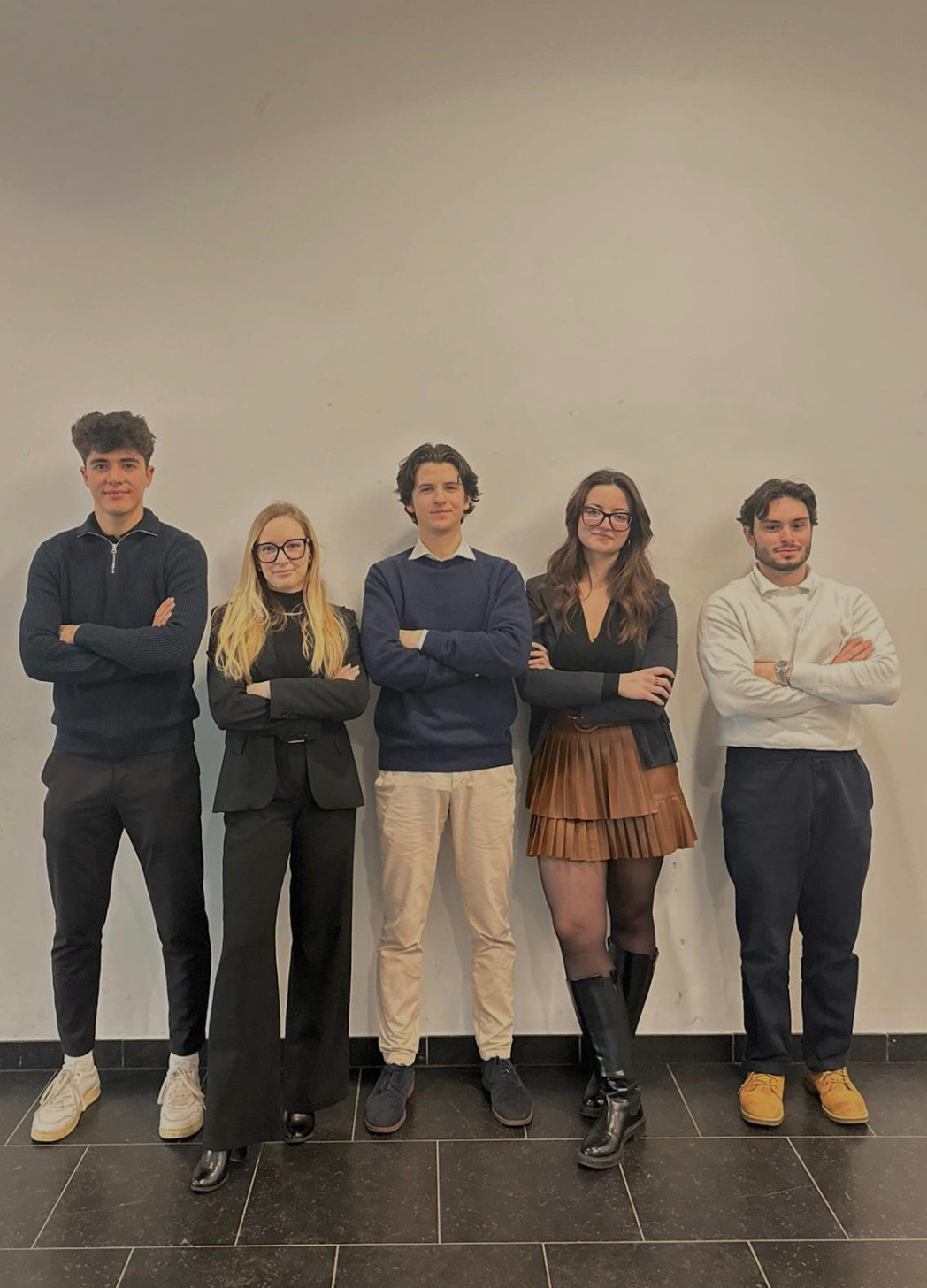 Five young adults stand side by side with arms crossed in front of a plain, light-colored indoor wall.