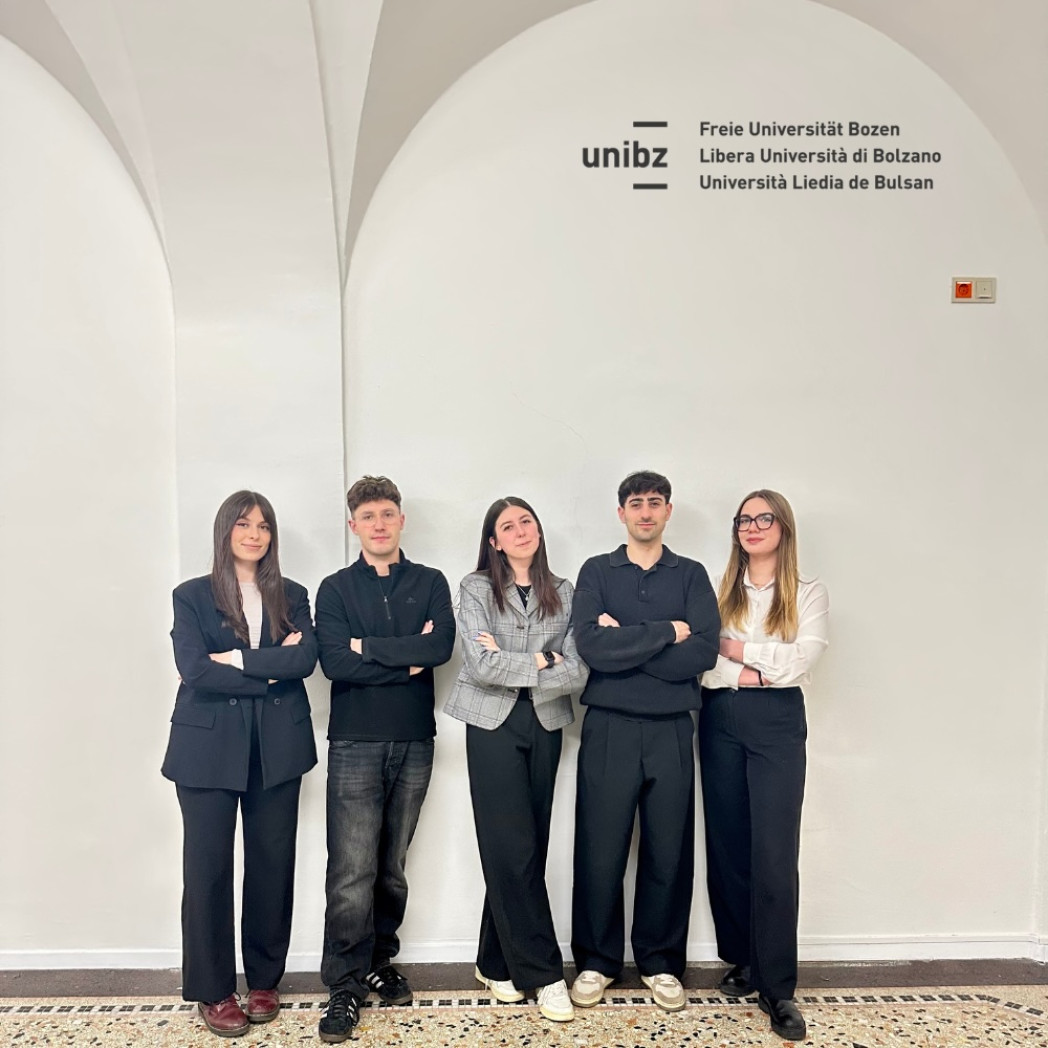 Five young adults stand side by side with arms crossed in front of a white wall displaying the Free University of Bozen-Bolzano (unibz) logo.