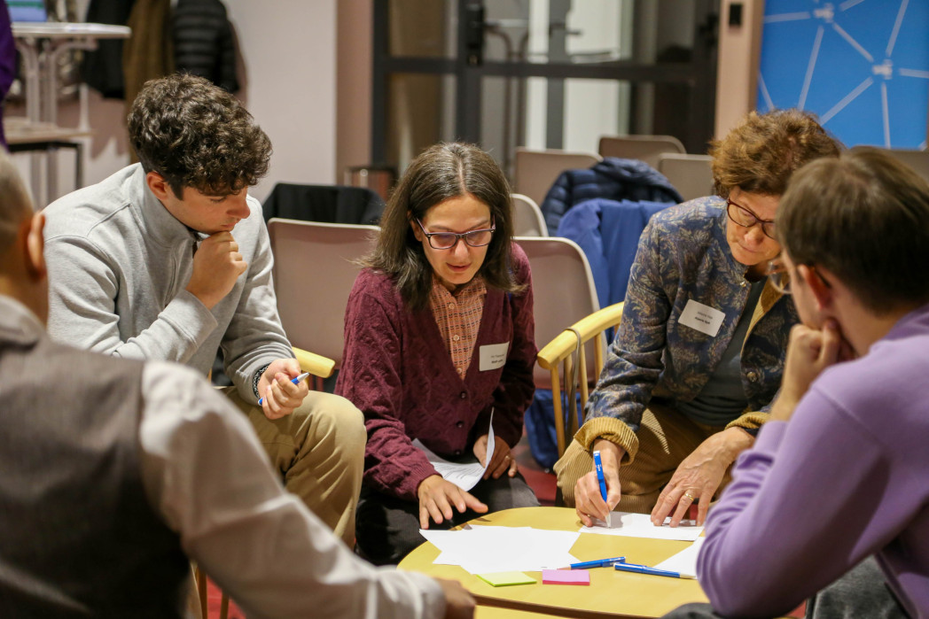Gruppe von Personen sitzt zusammen an einem Tisch, diskutiert und arbeitet konzentriert mit Notizen und farbigen Karten.