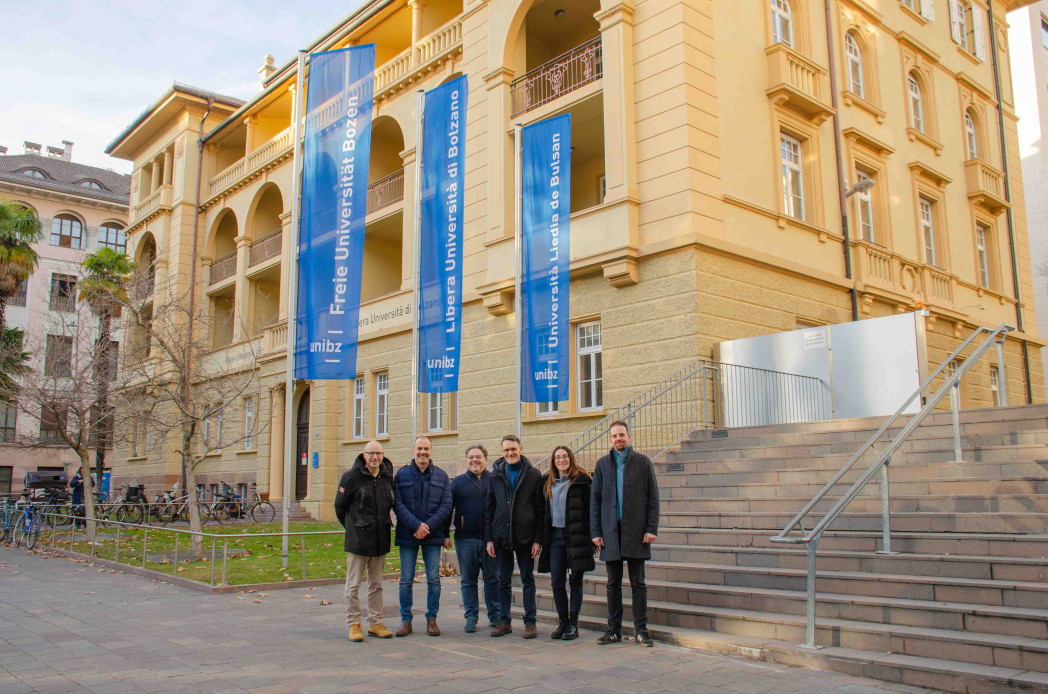 Sei persone davanti a un edificio di unibz. Dietro di loro sono appese tre bandiere della unibz. Accanto a loro una scalinata conduce verso l’alto.