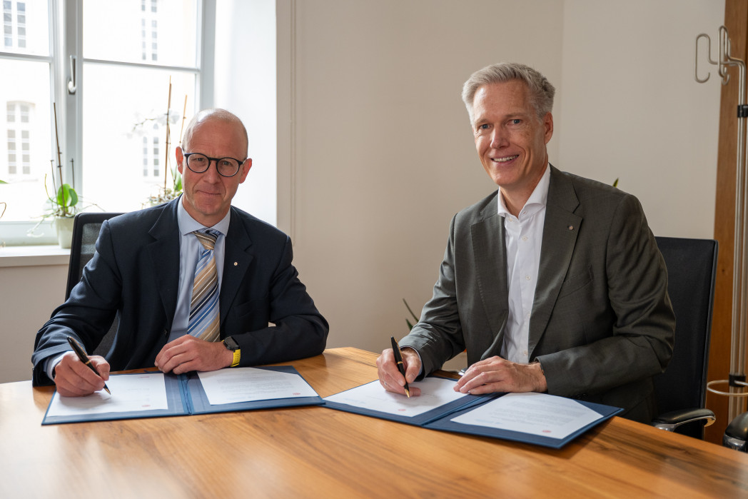 Two people are sitting at a table, signing a document placed in front of them