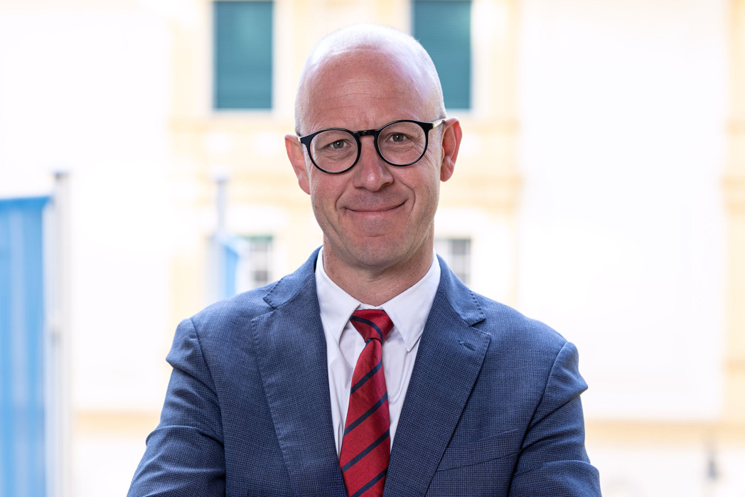 Porträtbild einer Person mit Brille und Glatze - mit Sakko und Krawatte; im Hintergrund ein unscharfes Gebäude.