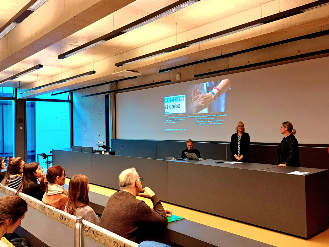 Ein Hörsaal. Auf der Leinwand ist die Präsentationsfolie mit dem Titel Connect zu sehen, während vorne drei Personen sprechen und moderieren.