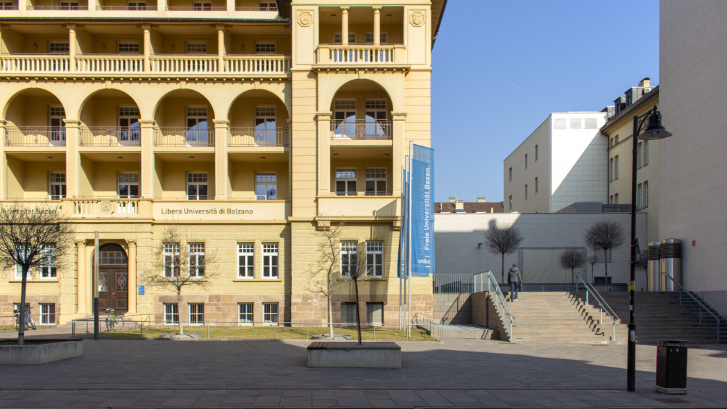 ellow historical building with arched columns and balconies facing an open plaza with modern white building adjacent