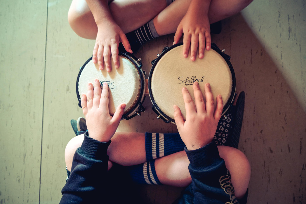 Zwei Kinder sitzen im Schneidersitz auf dem Boden. Zwischen ihnen eine Bongo-Trommel. In der Perspektive von oben sieht man ihre gekreuzten Beine und ihre Hände, die auf den Bongos.