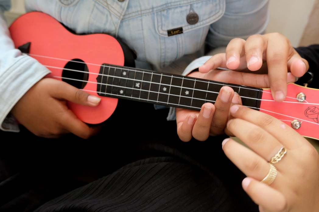 Vier Kinderhände auf einer Ukulele. Eine Kind zeigt dem anderen die Griffe.