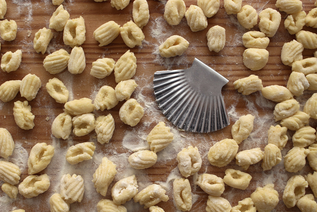 Gnocchibrett mit vielen grillten Gnocchi. In der Mitte liegt ein Küchenwerkzeug aus Edelstahl in Form einer Jakobsmuschel mit dem man die Gnocchi schneidet und die Rillen formt.