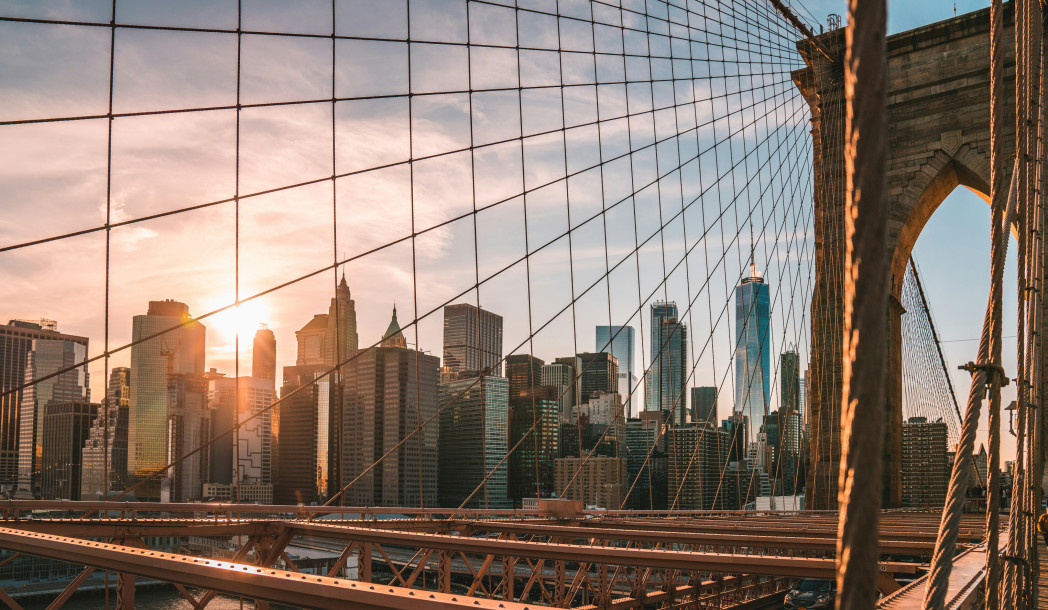 Vista dal Ponte di Brooklyn sullo skyline di New York al tramonto, con i cavi d'acciaio del ponte in primo piano.