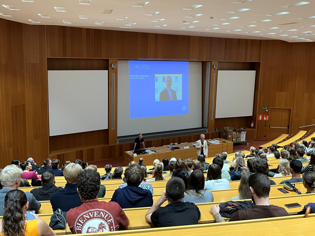 Students seated in tiered lecture hall watching presentation on central screen flanked by two blank projection screens.