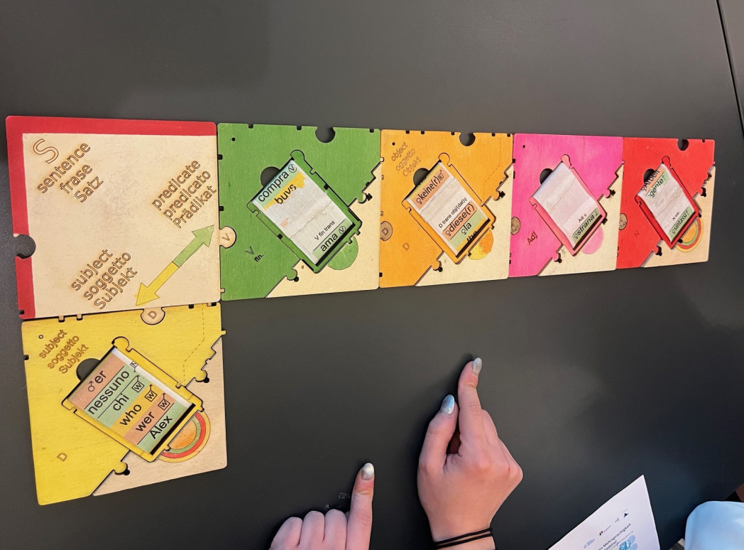 Ein buntes syntaktisches Puzzle mit sechs großen Puzzleteilen aus Holz auf einem Tisch, auf dem auch die Hände einer Person sichtbar sind.