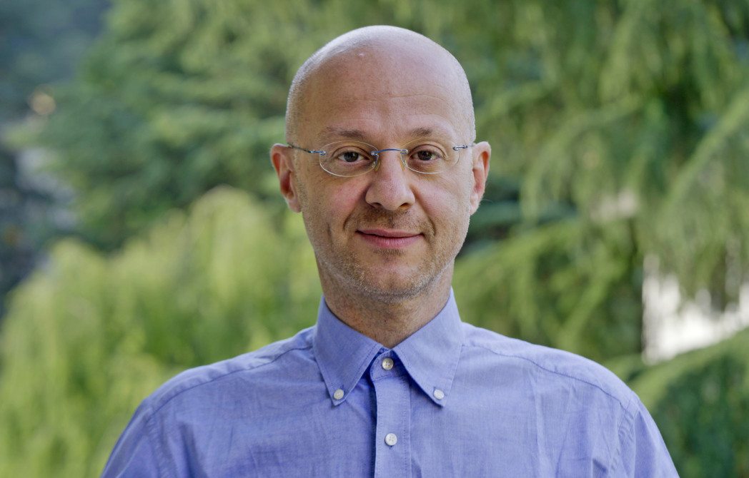 Un uomo con occhiali e camicia azzurra sorride leggermente, con alberi verdi sullo sfondo.