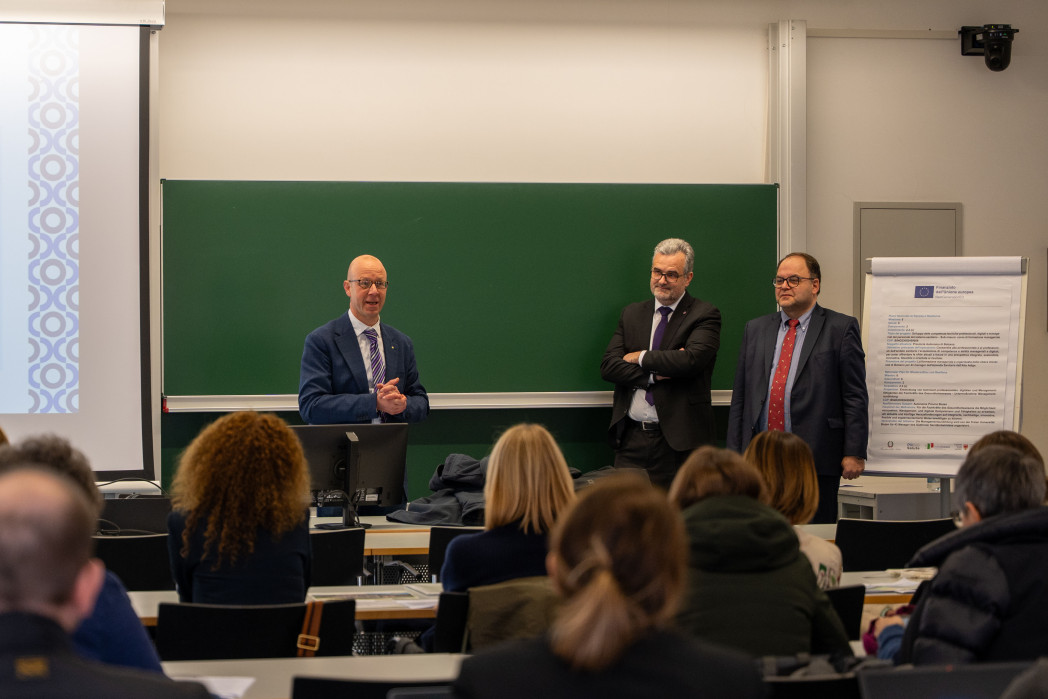 Tre uomini sono in piedi davanti a una lavagna verde in un’aula universitaria e si rivolgono a un pubblico seduto.