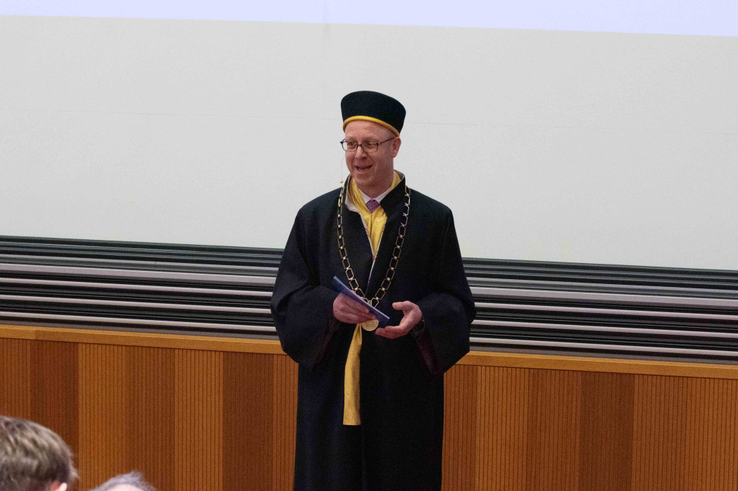 Eine Person in akademischer Robe mit Amtskette und schwarzer Kappe steht in einem Hörsaal und hält Notizen in den Händen. Im Hintergrund sind Holzvertäfelung und eine helle Projektionsfläche zu sehen.