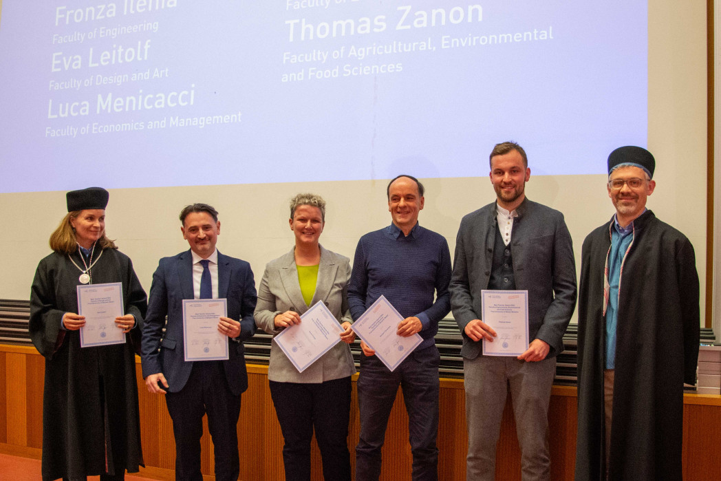 Sieben erwachsene Personen stehen nebeneinander auf einer Bühne in einem Hörsaal und halten Urkunden in den Händen. Sie tragen überwiegend formelle Kleidung; im Hintergrund ist eine Projektionsfläche mit Text zu sehen.