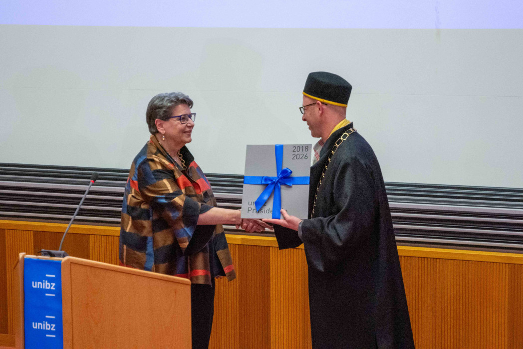 Zwei Personen stehen in einem Hörsaal und überreichen sich ein Buch mit blauer Schleife. Eine Person trägt eine akademische Robe mit Amtskette; im Hintergrund sind Holzvertäfelung und eine Projektionsfläche zu sehen.