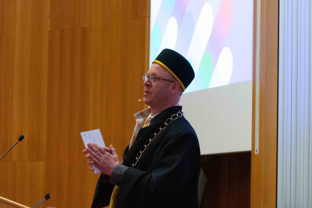 A person wearing an academic gown with a ceremonial chain and a cap speaks in a lecture hall, holding notes in their hands. A projection screen and wooden paneling are visible in the background.