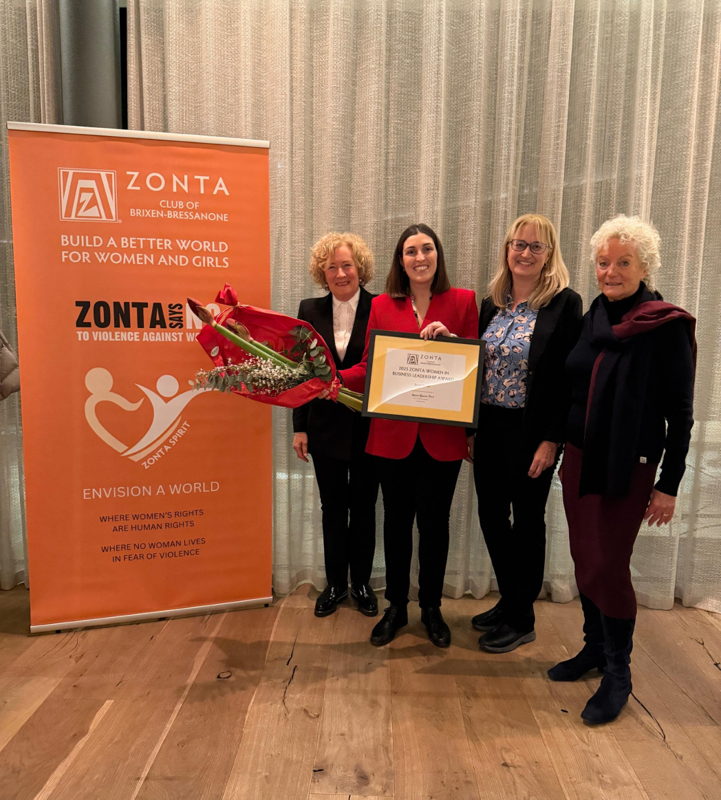 Fünf Frauen stehen nebeneinander vor einem Banner; eine Frau hält eine Urkunde und einen Blumenstrauß bei einer Veranstaltung.