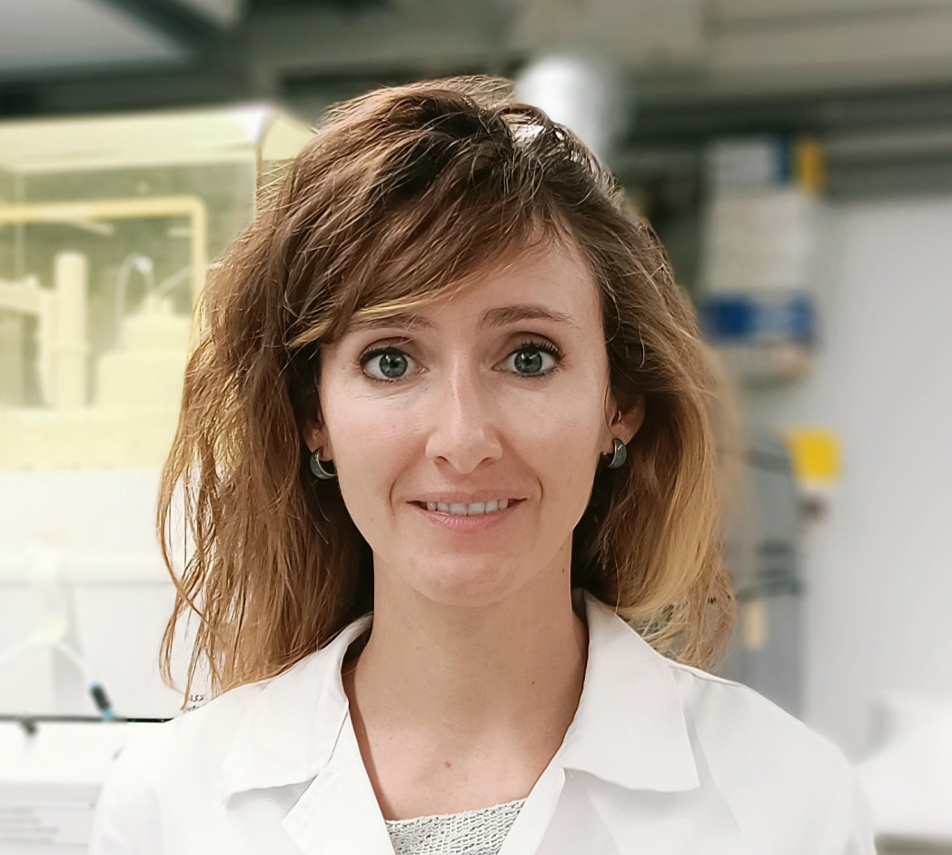 Donna sorridente in camice bianco da laboratorio guarda verso la camera, ambiente di laboratorio sfocato sullo sfondo.