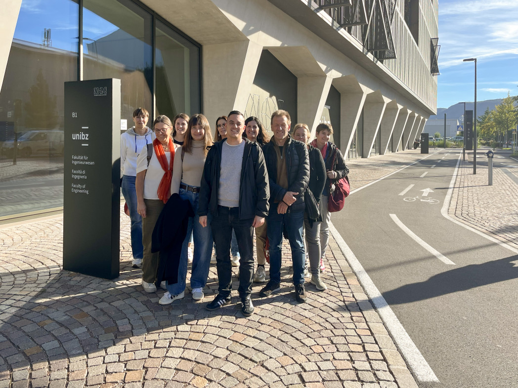 Gruppo di otto persone in piedi davanti a un edificio moderno con facciata in vetro in una giornata soleggiata