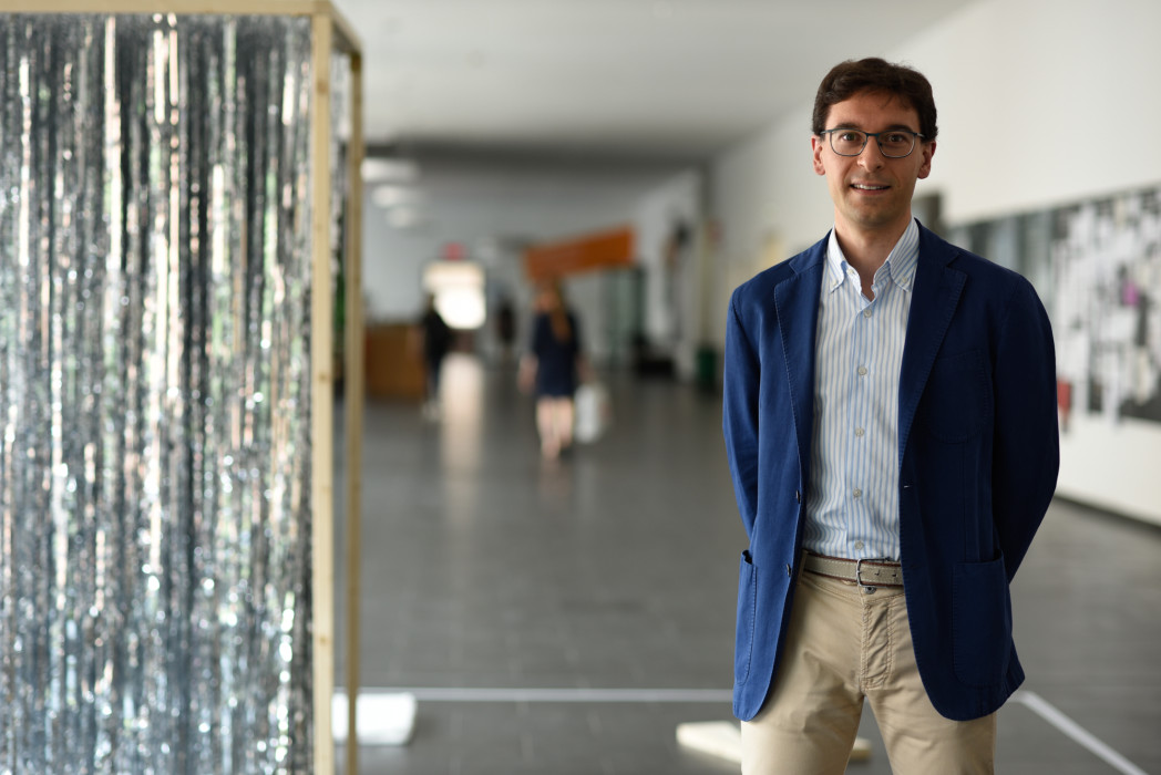 Uomo in piedi in un corridoio del campus, con giacca blu e occhiali, guarda verso la fotocamera; sullo sfondo decorazioni argentate e altre persone sfocate.