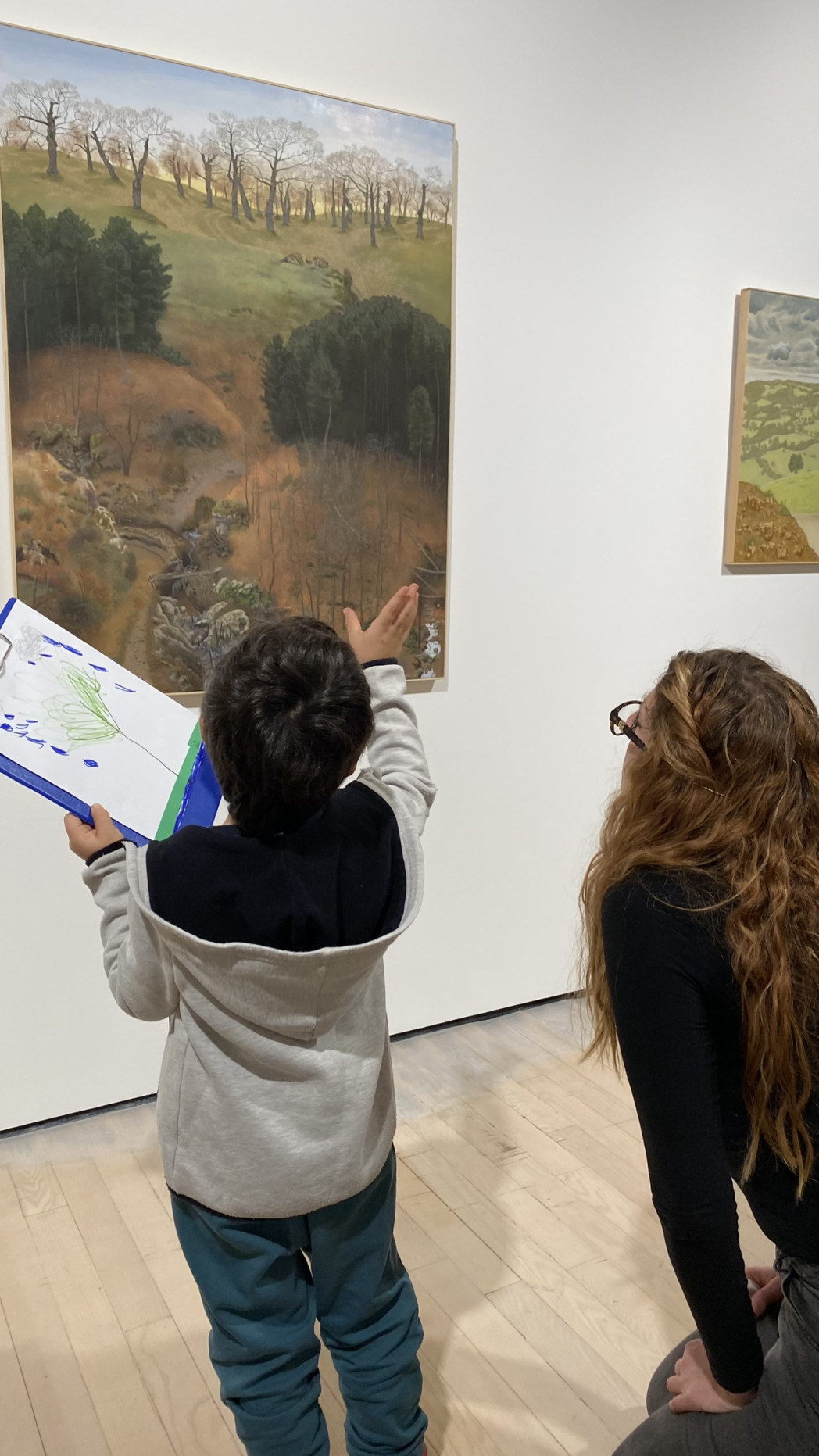 Bambino che indica un dipinto paesaggistico in un museo mentre una donna osserva accanto a lui con un foglio in mano.