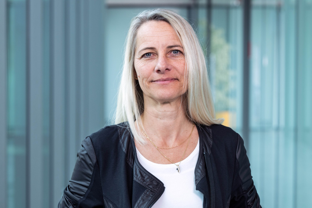 Ritratto di una donna con capelli fino alle spalle e espressione neutra, davanti a uno sfondo sfocato di finestre in vetro; indossa una giacca sopra una maglia e una collana sottile.