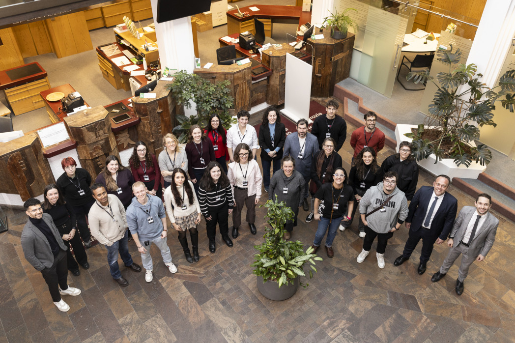 Gruppenfoto von etwa 25 Personen mit Namensschildern, die in einem halbkreisförmigen Foyer mit Holztheken und Pflanzen stehen und in die Kamera lächeln.
