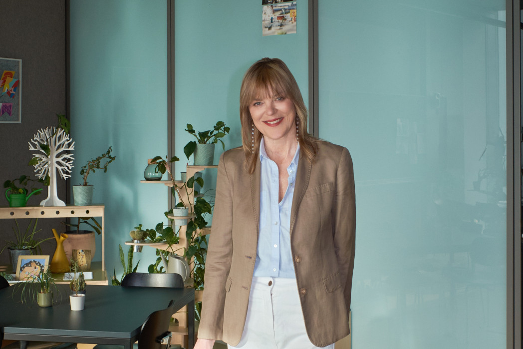 Ritratto di una donna sorridente con capelli fino alle spalle, in piedi in un ufficio luminoso davanti a una parete in vetro; indossa un blazer e una camicia chiara, sullo sfondo si vedono piante da interno e un tavolo.