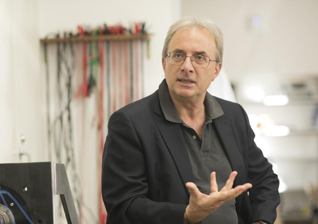 Uomo parla gesticolando, in un ambiente tecnico con cavi e strumenti sullo sfondo.