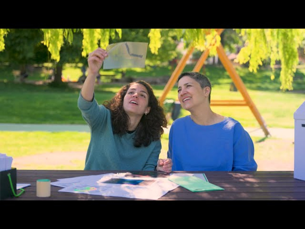 Two women are sitting at a table in a park looking at a picture that one of them is holding up to the light.