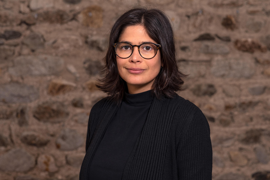 Porträt einer Frau mit Brille und dunkler Kleidung vor einer Natursteinwand; sie blickt ruhig in die Kamera.