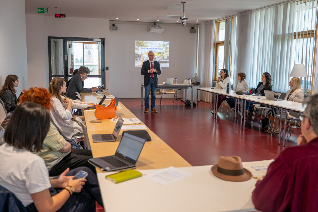 A man gives a presentation to a group of people seated around a U-shaped table in a bright conference room.