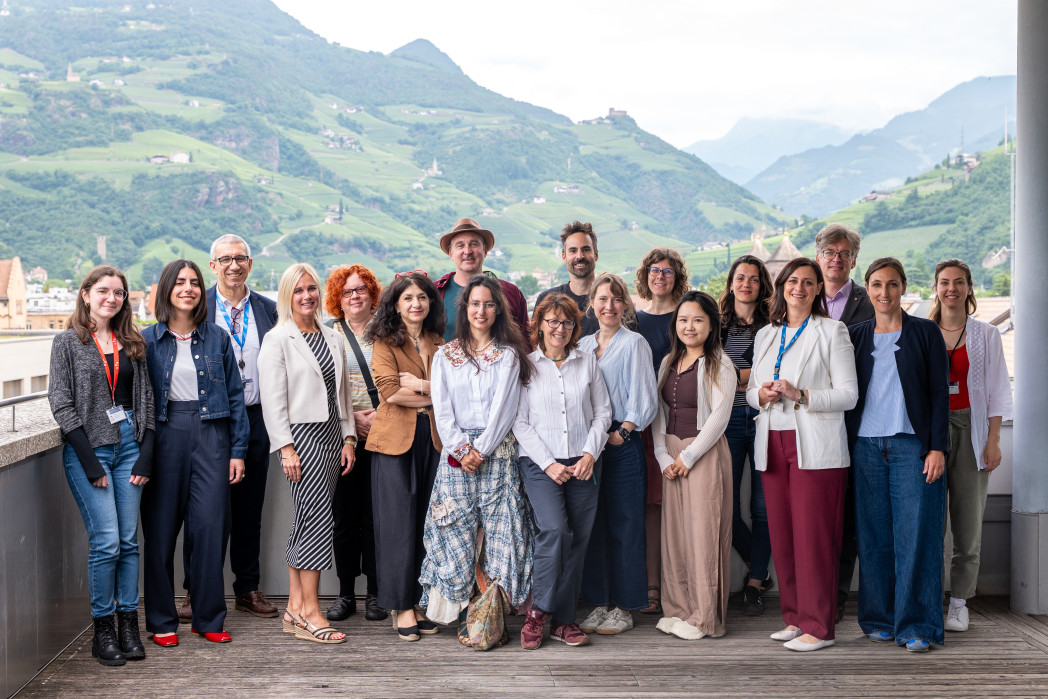 A group of people poses for a photo on a terrace with a scenic mountain landscape in the background.