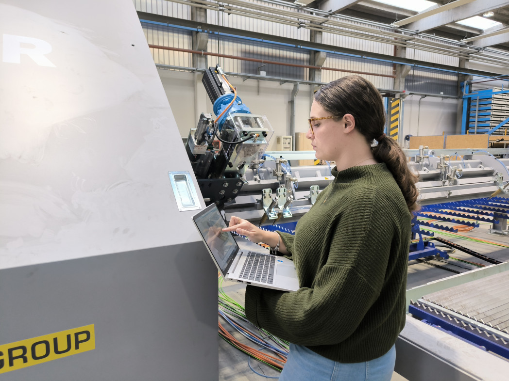 Frau bedient einen Laptop an einer Maschine. Sie steht in einer industriellen Halle.