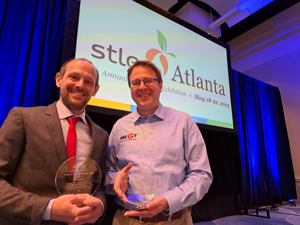 Two men in a hall; one is holding an award. Behind them, a projection screen.