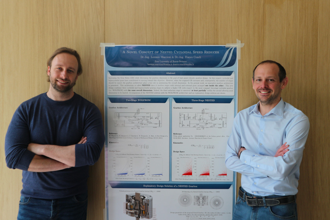 Two men, Lorenzo Maccioni and Franco Concli, standing in front of a wall; between them, a poster is displayed.