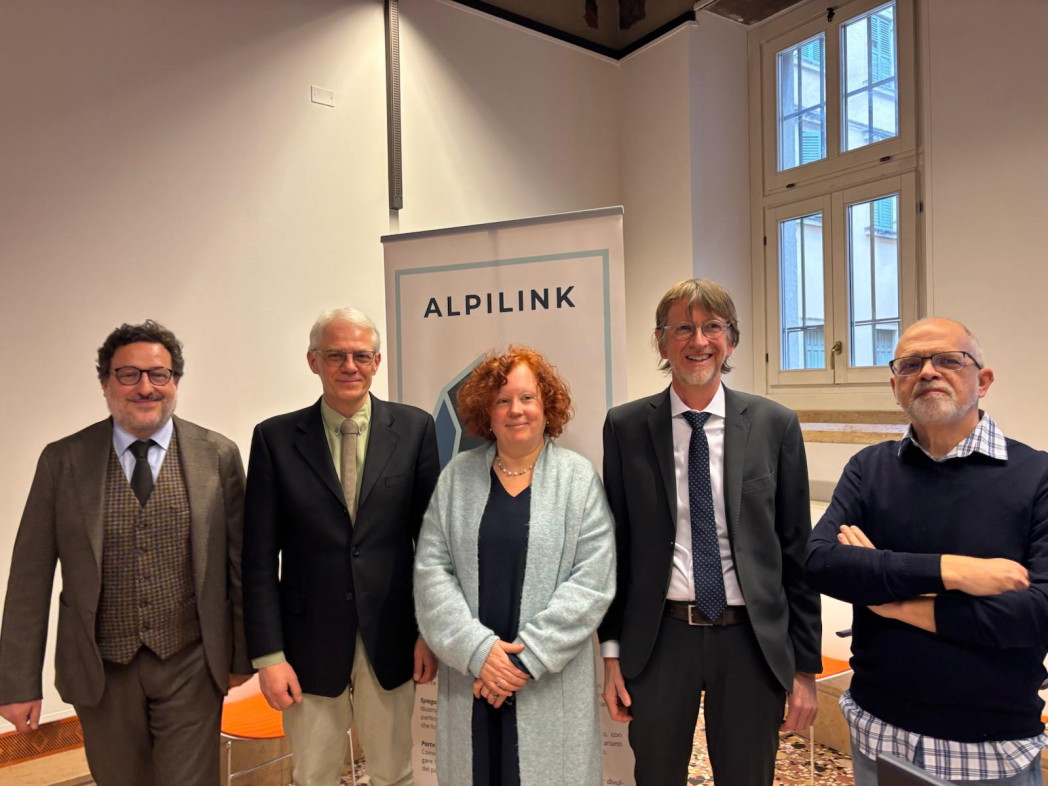 Fünf Personen stehen in einem Raum nebeneinander; im Hintergrund ein Roll-Up mit der Aufschrift „Alpilink“.