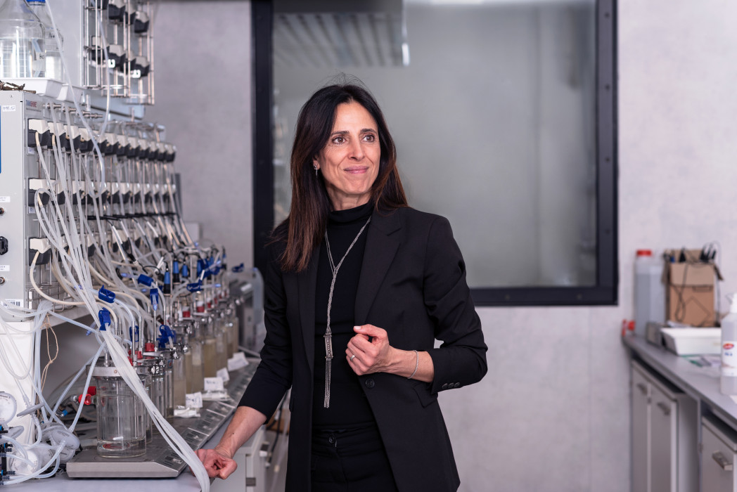 Una donna si trova in un laboratorio accanto a un’apparecchiatura complessa con tubi e contenitori di vetro, e guarda attentamente di lato.